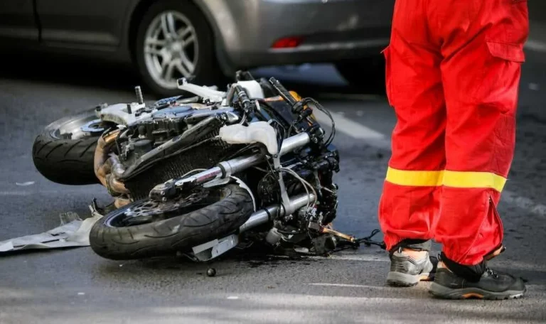 Wrecked motorcycle at an accident scene in New York City with a first responder in a red uniform standing nearby