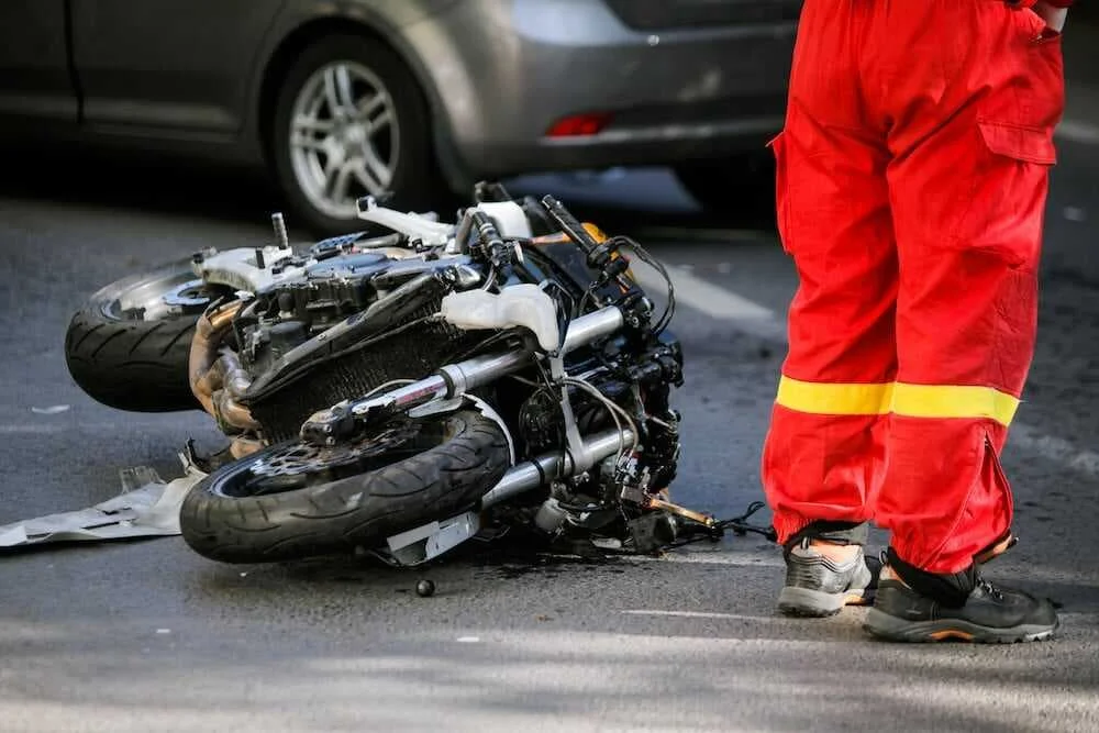 Wrecked motorcycle at an accident scene in New York City with a first responder in a red uniform standing nearby
