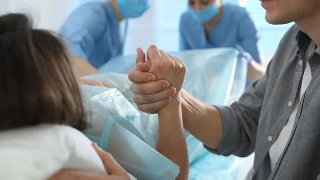 Husband holding his wife's hand during childbirth as medical staff assist in the delivery room — birth injury lawyer NYC